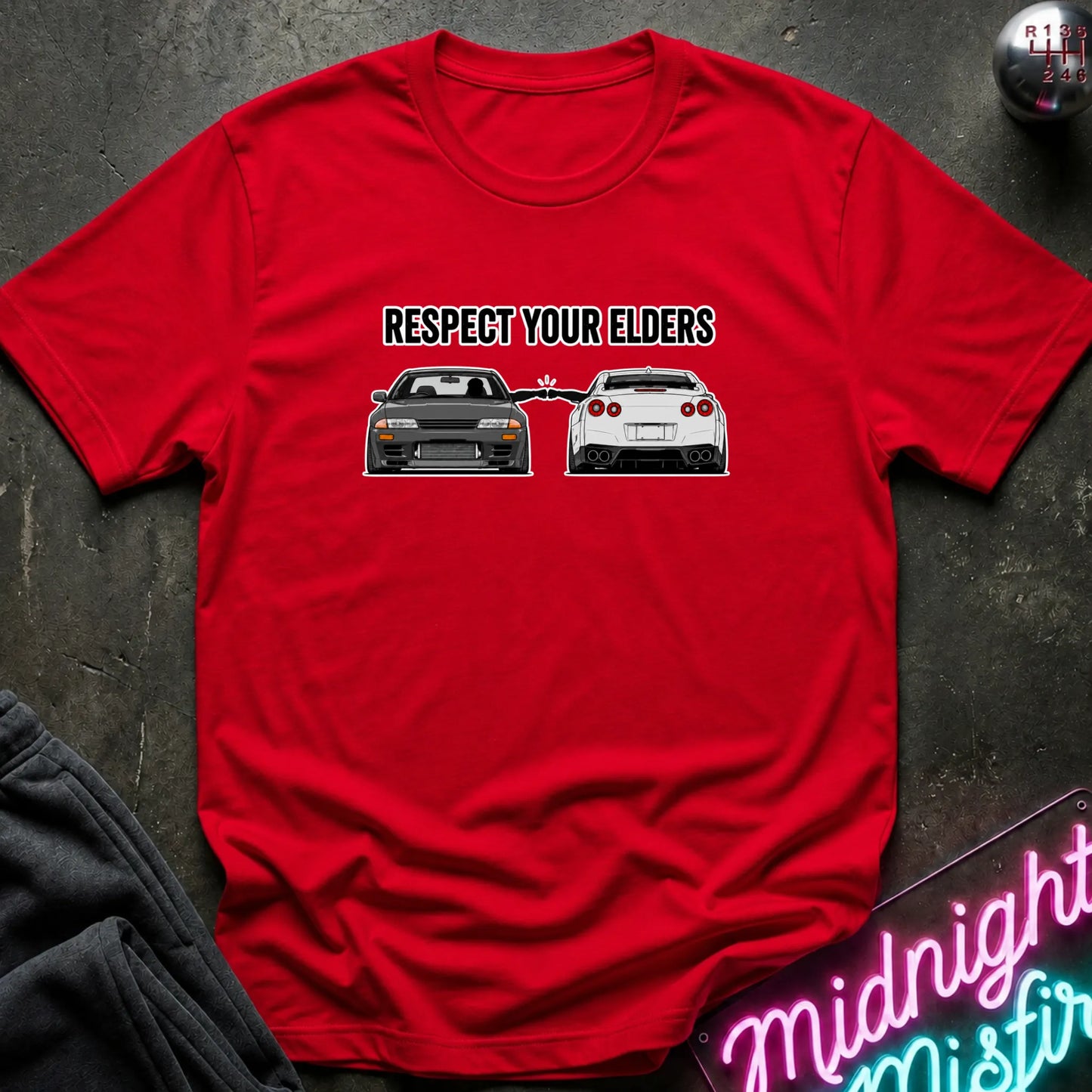 A red short-sleeve graphic T-shirt laid flat on a dark textured surface. The shirt has a black printed slogan reading 'RESPECT YOUR ELDERS' above two cars, with a neon sign and dark clothing partially visible around it.