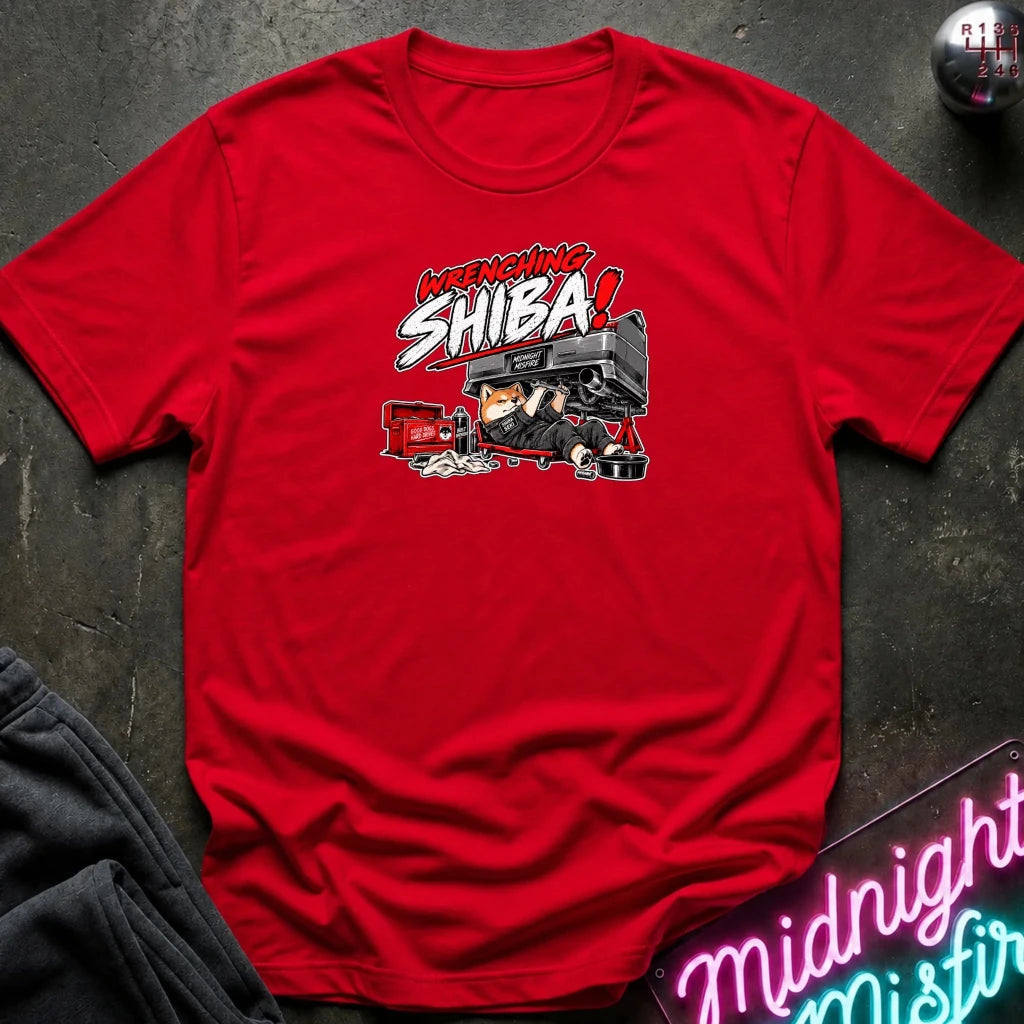 A red short-sleeve graphic t-shirt laid flat on a dark textured surface. The graphic features the text 'WRENCHING SHIBA!' in red and white comic-style lettering above an illustration of a character in a black and red vehicle.