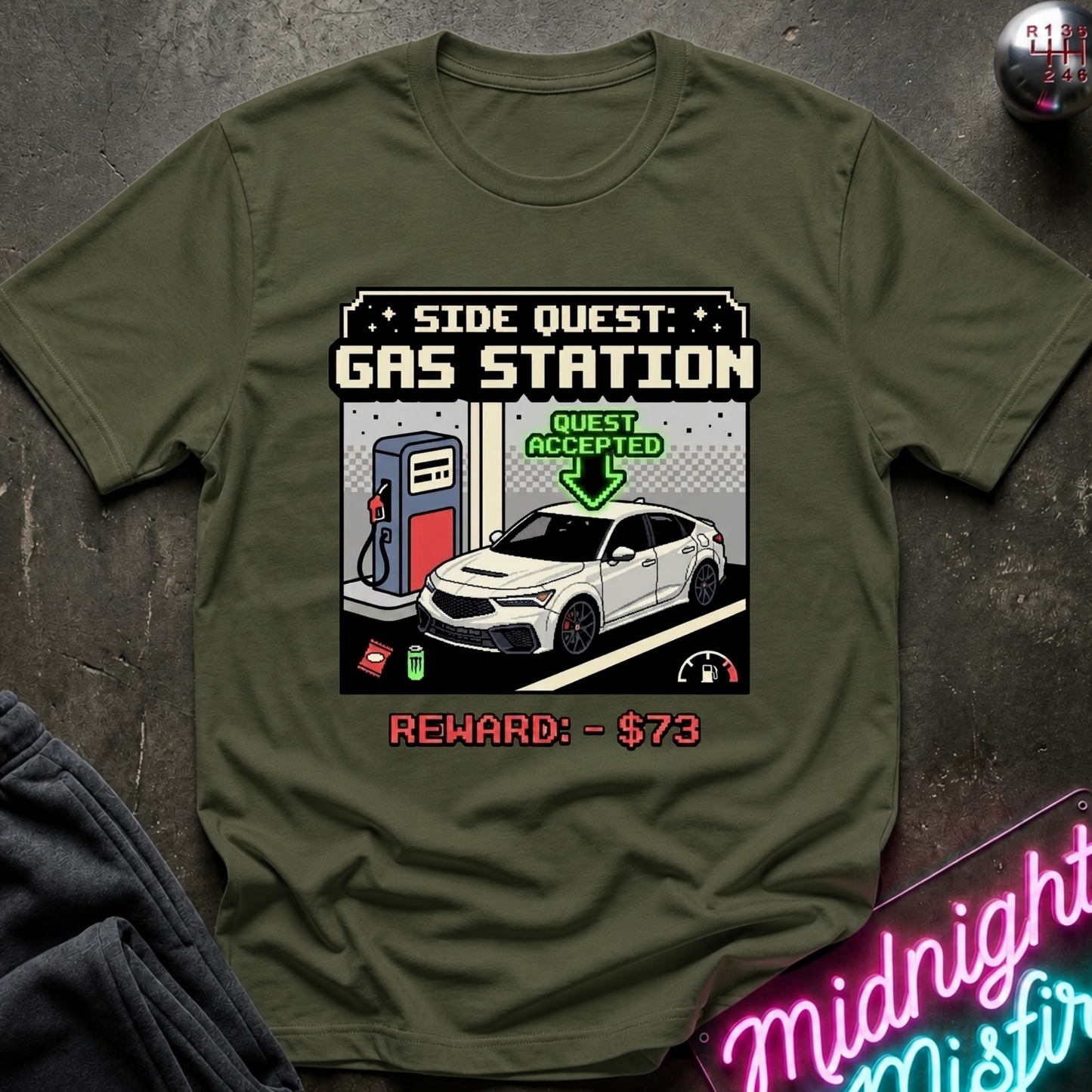 An olive green short-sleeve t-shirt with a 'Side Quest: Gas Station' graphic print, laid flat on a dark textured surface with a neon sign and dark fabric in the corners.
