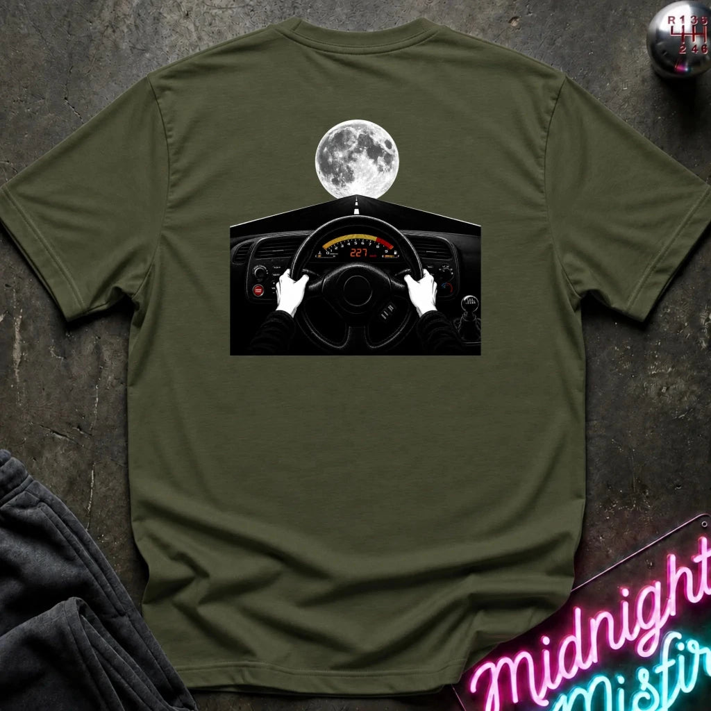 An olive green short-sleeve t-shirt laid flat on a dark textured surface, featuring a graphic print of a car dashboard and steering wheel with a full moon above. 