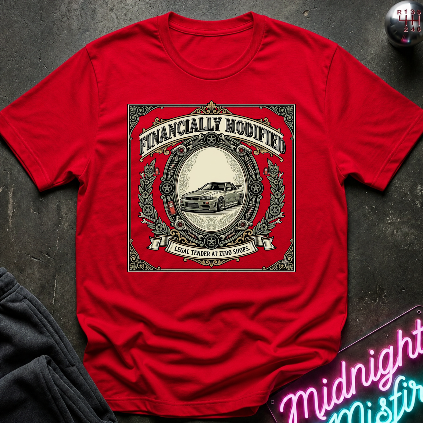 A red short-sleeve graphic t-shirt laid flat on a dark textured surface. The shirt features a large vintage-style circular graphic with the text 'FINANCALLY MODIFIED' and 'LOCAL TENDER AT ZERO SHOPS' surrounding an illustration of a classic car.