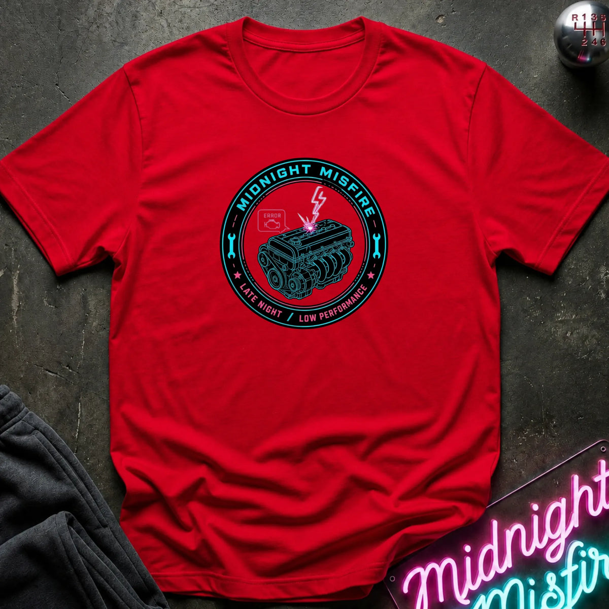 A red short-sleeve crewneck t-shirt laid flat on a dark textured surface, featuring a circular teal and black graphic on the chest that reads 'Midnight Misfire' with an engine illustration and lightning bolt. A neon sign and folded dark fabric are partially visible around the shirt.