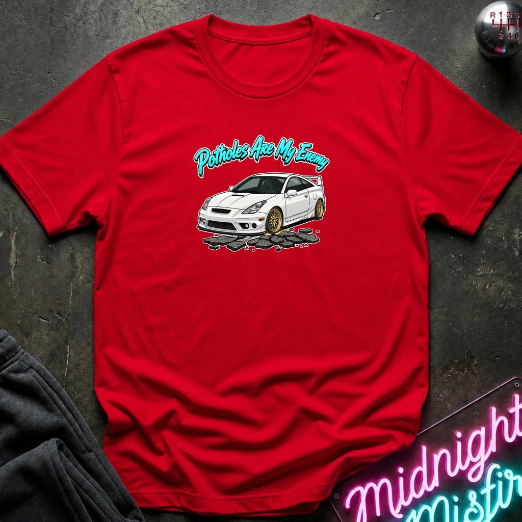 A red short-sleeve t-shirt laid flat on a dark textured surface, featuring a graphic of a white sports car with gold wheels and the text 'Pistons Are My Enemy' in teal and white. 