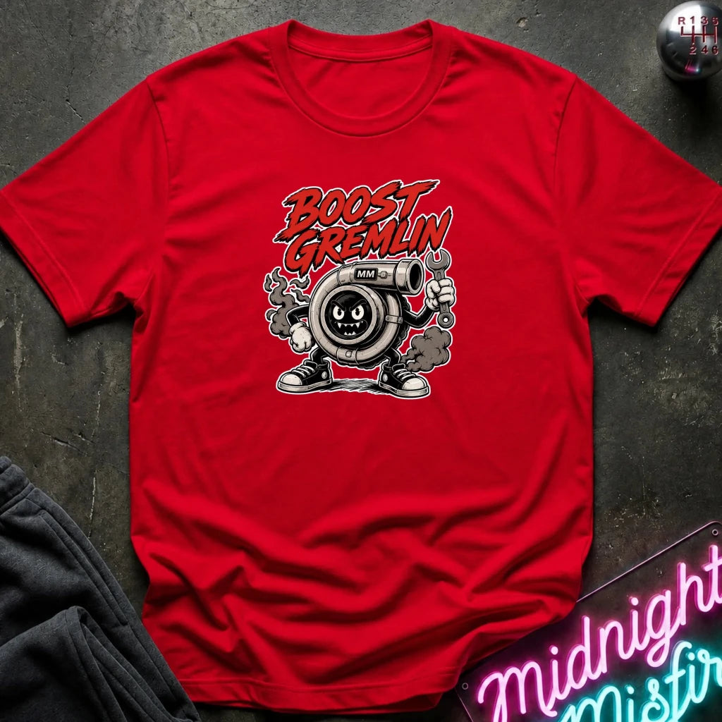 A red short-sleeve graphic T-shirt laid flat on a dark textured surface. The shirt features a cartoon graphic of a gas gauge character with the text 'BOOST GREMLIN' in red and black above it.