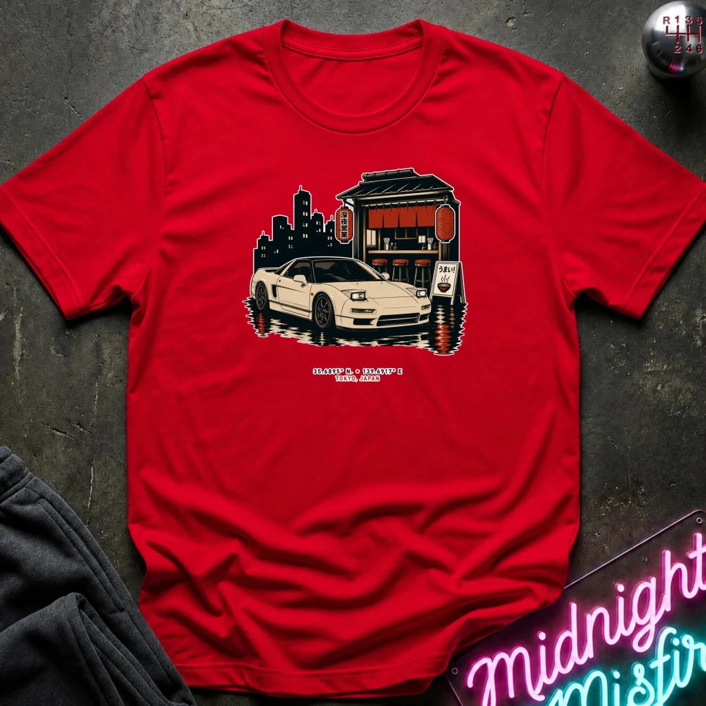 A red short-sleeve crewneck t-shirt laid flat on a dark textured surface, featuring a graphic print of a white convertible sports car parked in front of a Japanese-style building and city skyline. The shirt is styled with a dark gray sweatshirt and a neon sign in the background.