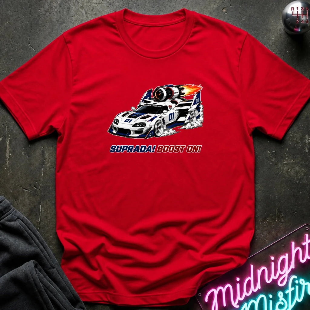 A red short-sleeve crewneck t-shirt featuring a graphic of a white and blue Supra race car with flames and the text 'SUPRADA! BOOST ON!' printed on the chest. The shirt is laid flat on a dark textured surface with a gray sweatshirt and a neon sign partially visible.