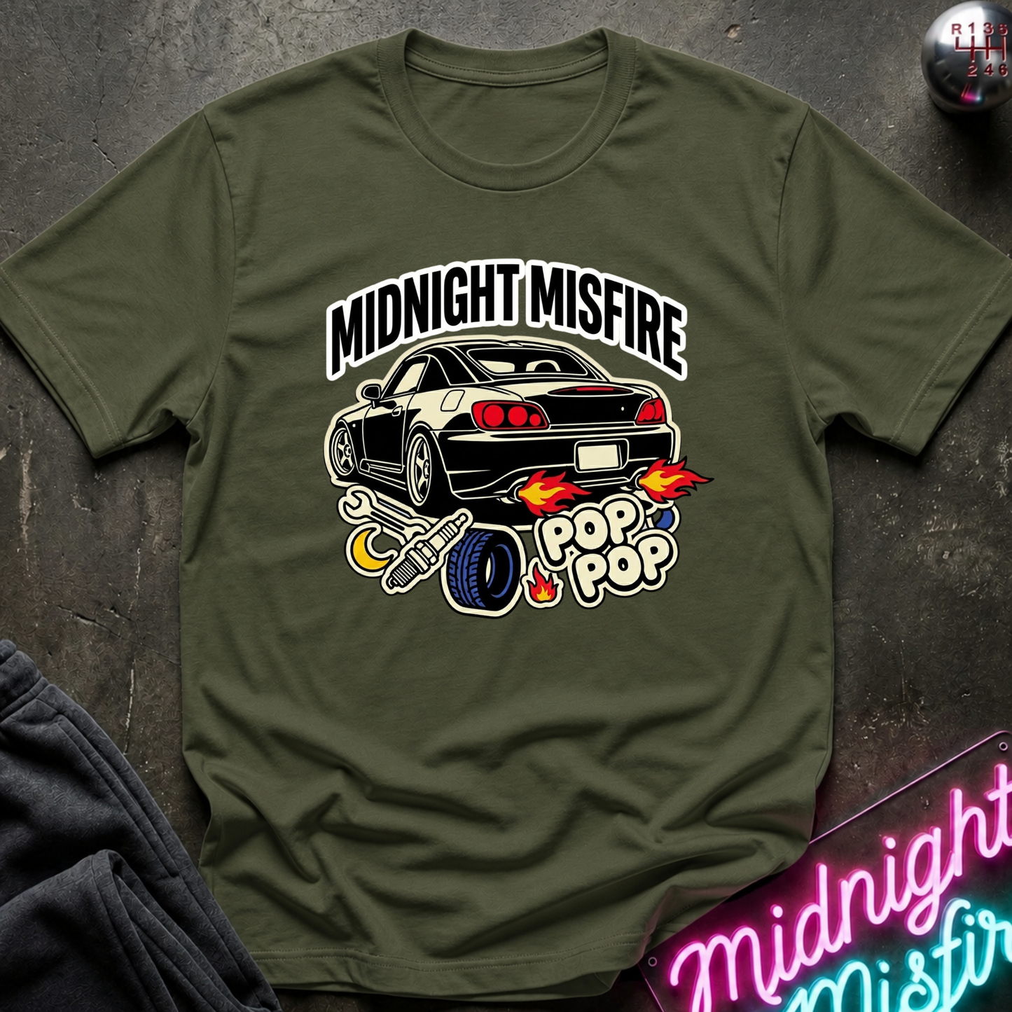 An olive green short-sleeve t-shirt with a graphic of a black sports car and flames, with the text 'MIDNIGHT MISFIRE' and 'POP POP', laid flat on a dark textured surface.