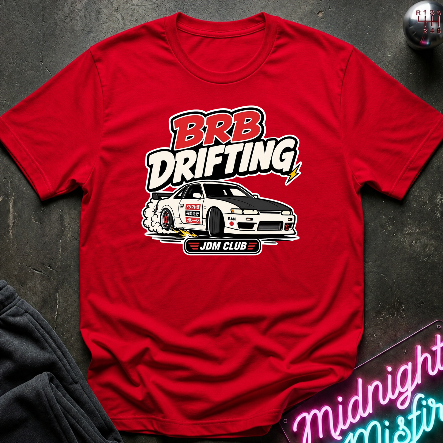 A red short-sleeve graphic T-shirt laid flat on a dark textured surface. The shirt features a large front print reading “BRB Drifting” above a white drifting car illustration and the text “JDM CLUB.