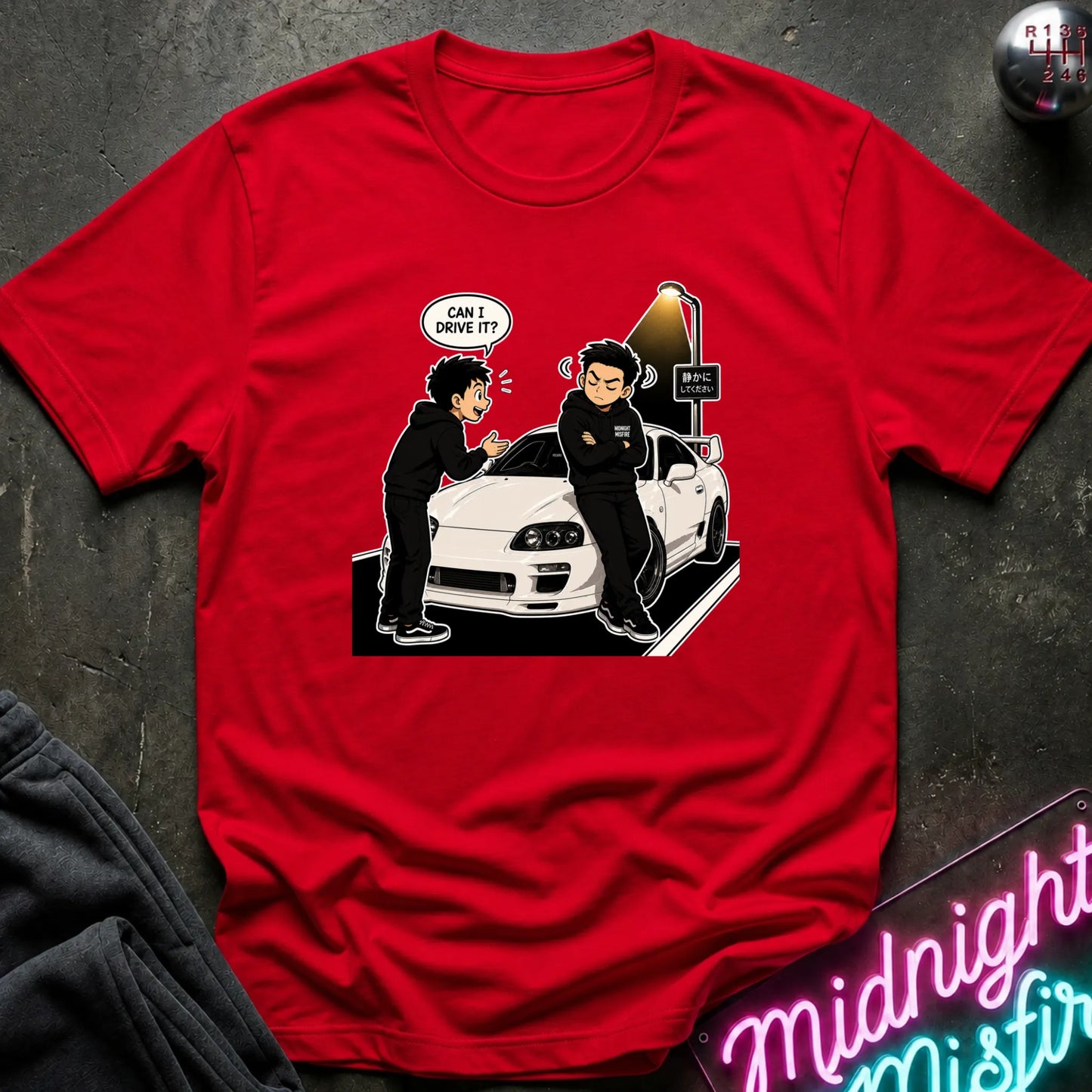 A red short-sleeve crewneck t-shirt featuring a cartoon graphic of two men in black standing by a white sports car, with one speech bubble reading 'CAN I DRIVE IT?'. The shirt is laid flat on a dark textured surface with a gray sweatshirt corner and a neon sign visible as props.