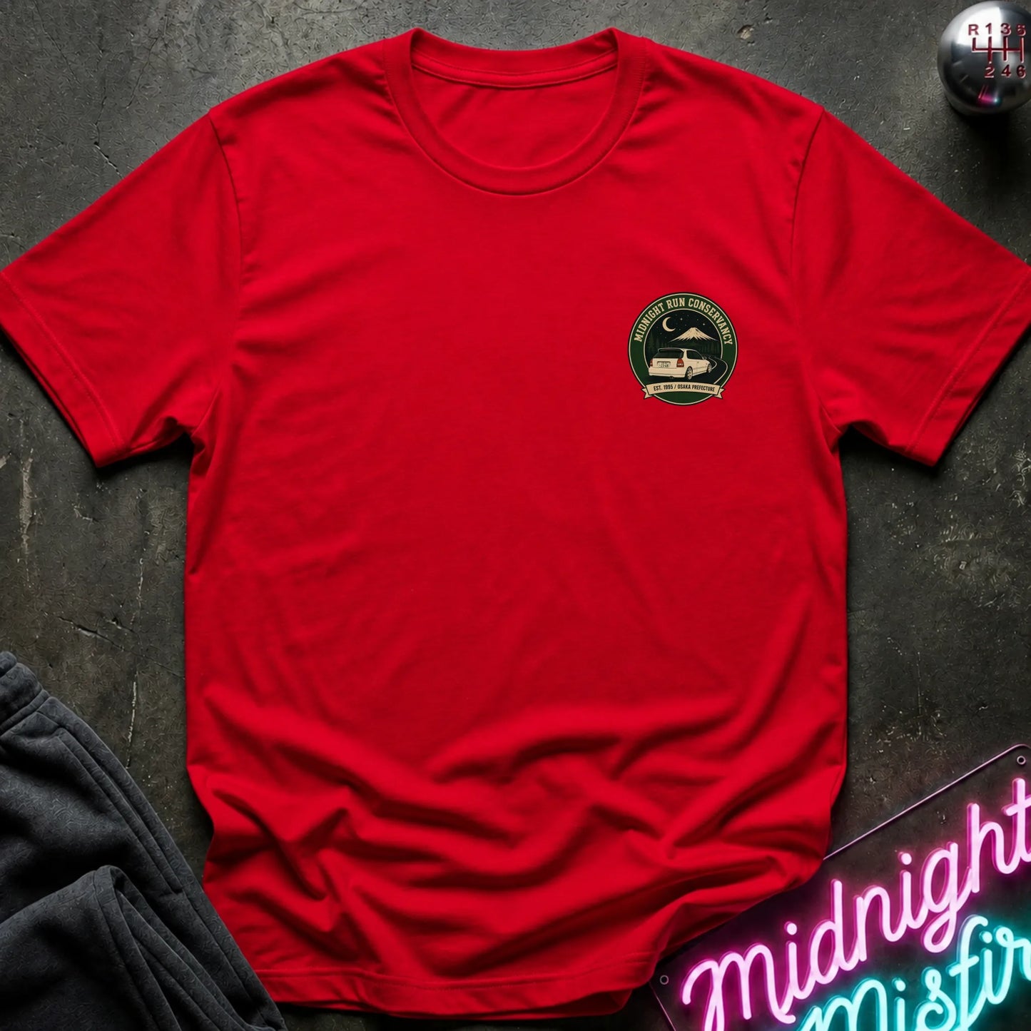 A red short-sleeve crewneck t-shirt with a circular green and black chest graphic, styled flat-lay on a dark textured surface with a neon sign and dark pants nearby.