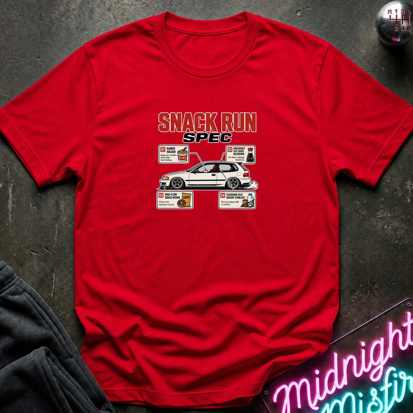 A red short-sleeve graphic t-shirt laid flat on a dark textured surface. The graphic features the text 'SNACK RUN SPEC' above a white car, surrounded by small food and drink illustrations.