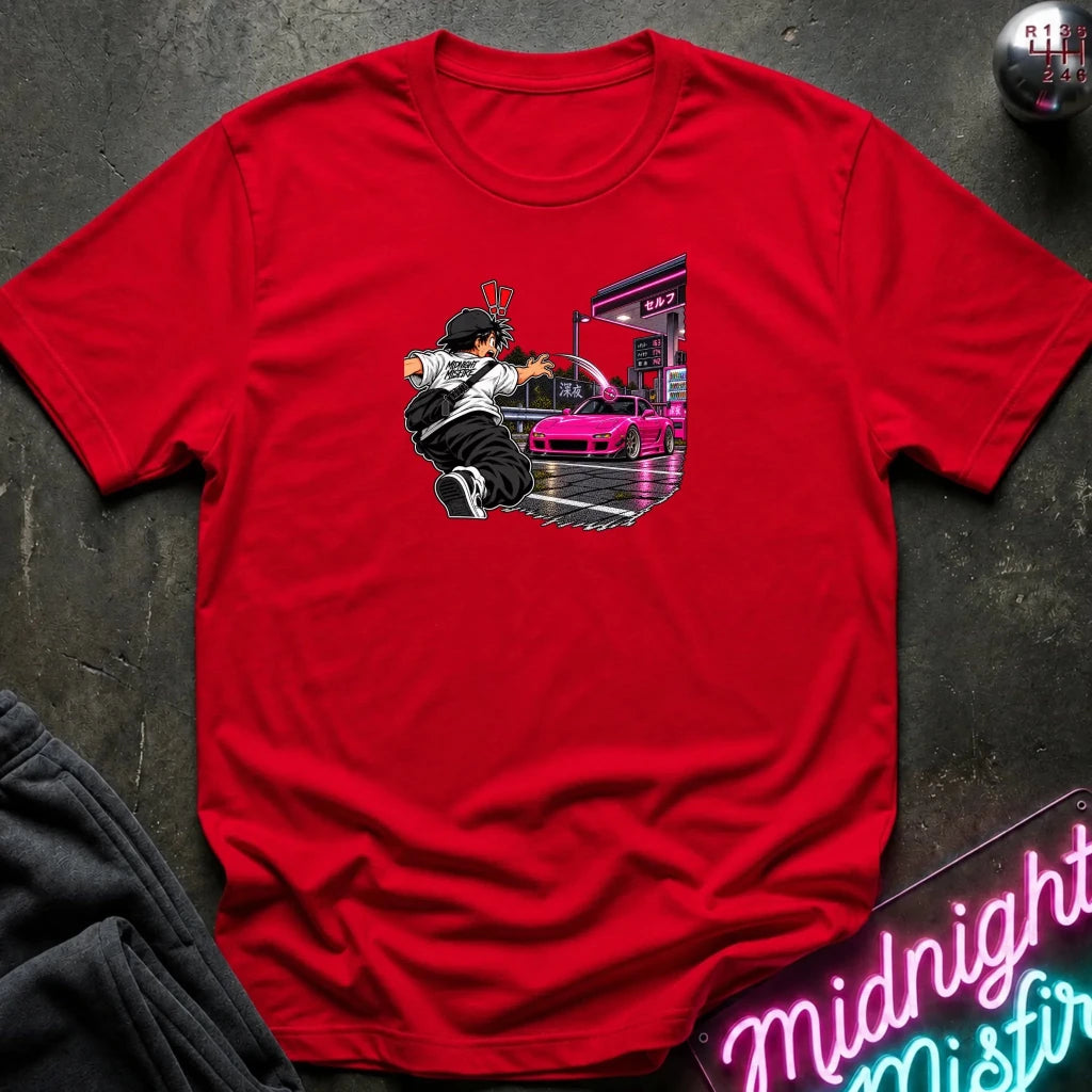 A red short-sleeve crewneck t-shirt laid flat on a dark textured surface. The center of the shirt features a graphic print of an anime character in a black and white outfit with a hat, next to a sports car 