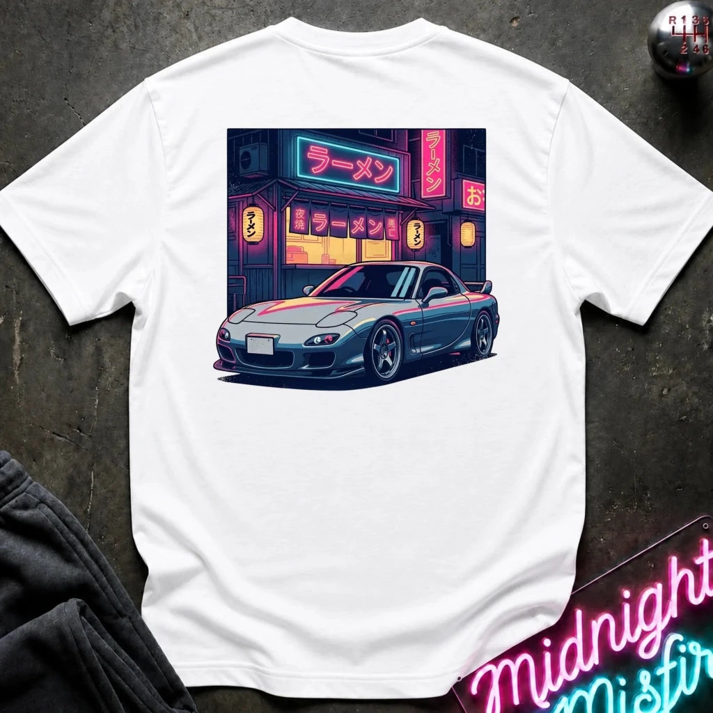 A white short-sleeve t-shirt laid flat on a dark textured surface, featuring a colorful graphic print of a blue sports car parked in front of a neon-lit ramen shop.
