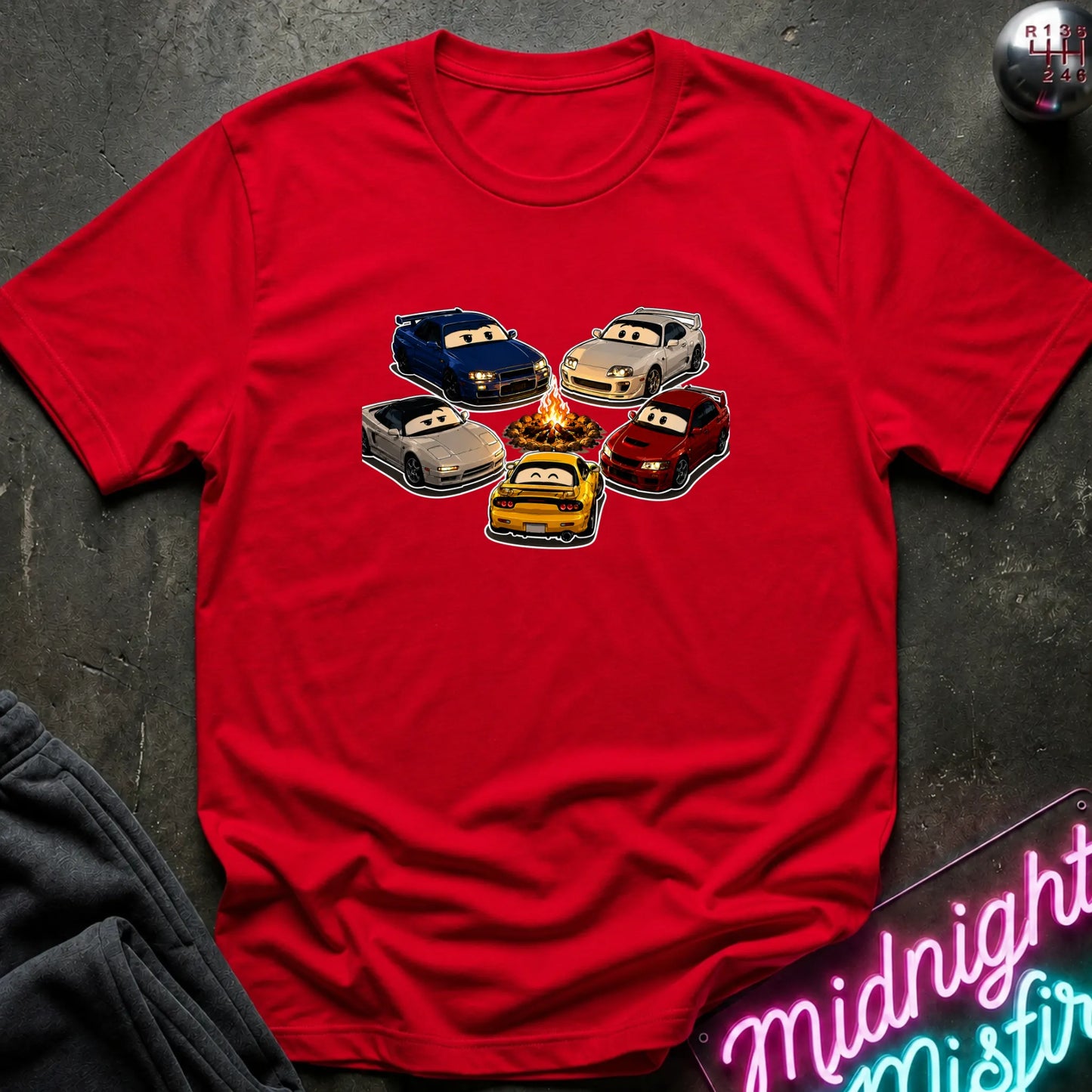 A red short-sleeve t-shirt laid flat on a dark textured surface, featuring a graphic of cartoon race cars arranged around a campfire. The shirt is styled with a gray sweatshirt in the corner and a pink neon sign in the bottom right.