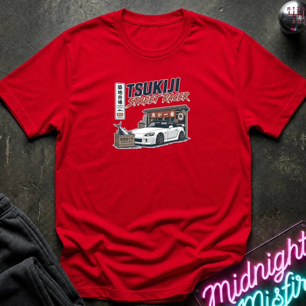 A red short-sleeve crewneck t-shirt featuring a graphic print of a white Nissan Sentra R13 with 'TSUKIJI STREET RICKER' text, styled flat on a dark textured background with a neon sign and dark pants partially visible.