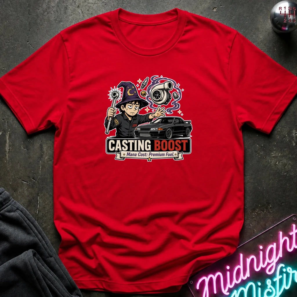A red short-sleeve graphic t-shirt laid flat on a dark textured surface. The graphic features a cartoon wizard holding a wand next to a black car, with the text 'CASTING BOOST' 
