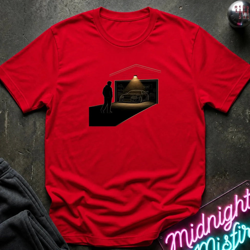 A red short-sleeve crewneck t-shirt laid flat on a dark textured surface. The center of the shirt features a graphic print of a silhouette of a man standing in a dark room with a car parked inside under a spotlight, with a garage door opening above.
