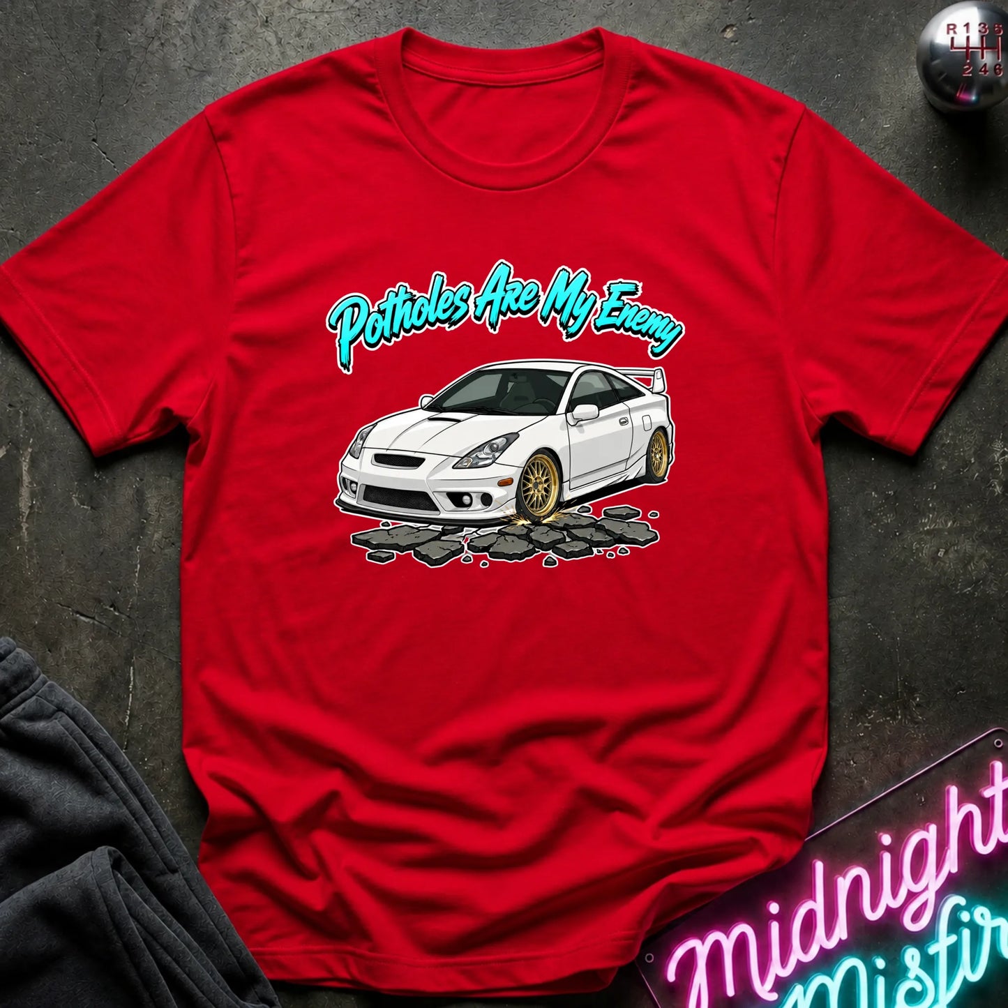 A red short-sleeve graphic T-shirt laid flat on a dark textured surface. The shirt features a white car illustration with gold wheels and the phrase 'Potatoes Are My Enemy' in teal script above it.