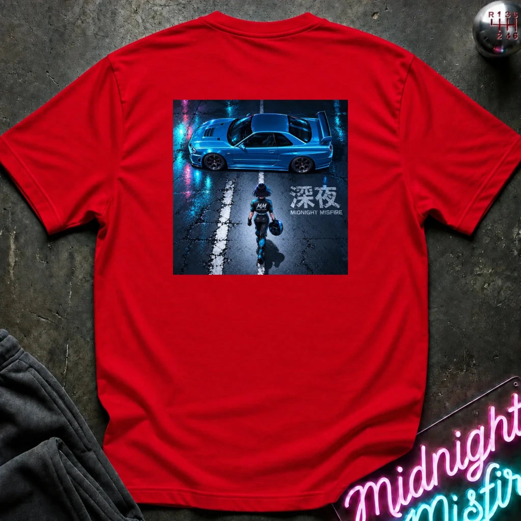A bright red short-sleeve t-shirt laid flat on a dark textured surface, featuring a large square graphic print on the back with a blue car at night and a person walking in the street. The shirt is styled with a gray sweatshirt, a gear shift knob, and a neon sign in the background.