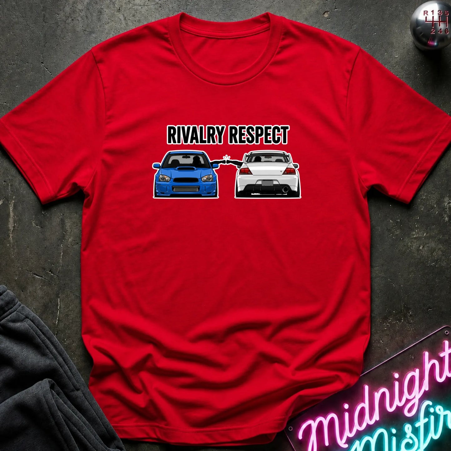 A red short-sleeve graphic t-shirt laid flat on a dark textured surface. The graphic features the text 'RIVALRY RESPECT' above a blue and white car, 