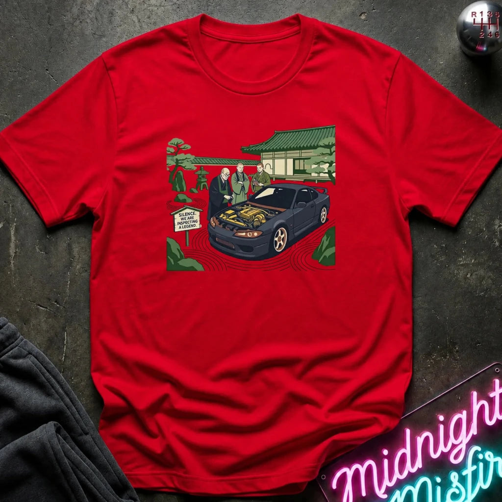 A bright red short-sleeve crewneck t-shirt laid flat on a dark textured surface. The center of the shirt features a detailed graphic print of a dark blue sports car with its hood open, surrounded by three men in suits, a Japanese-style building, and green trees.