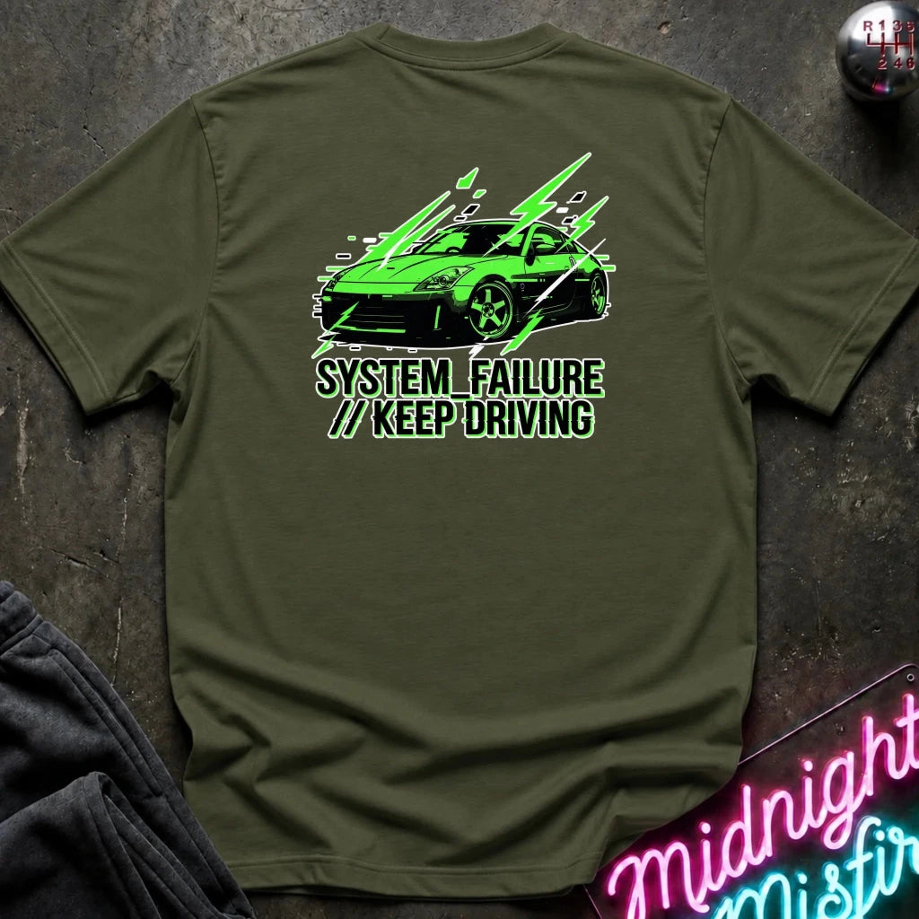 An olive green short-sleeve t-shirt laid flat on a dark textured surface, featuring a graphic of a green sports car with text 'SYSTEM FAILURE // KEEP DRIVING' in green. A neon sign and dark clothing are partially visible as props.