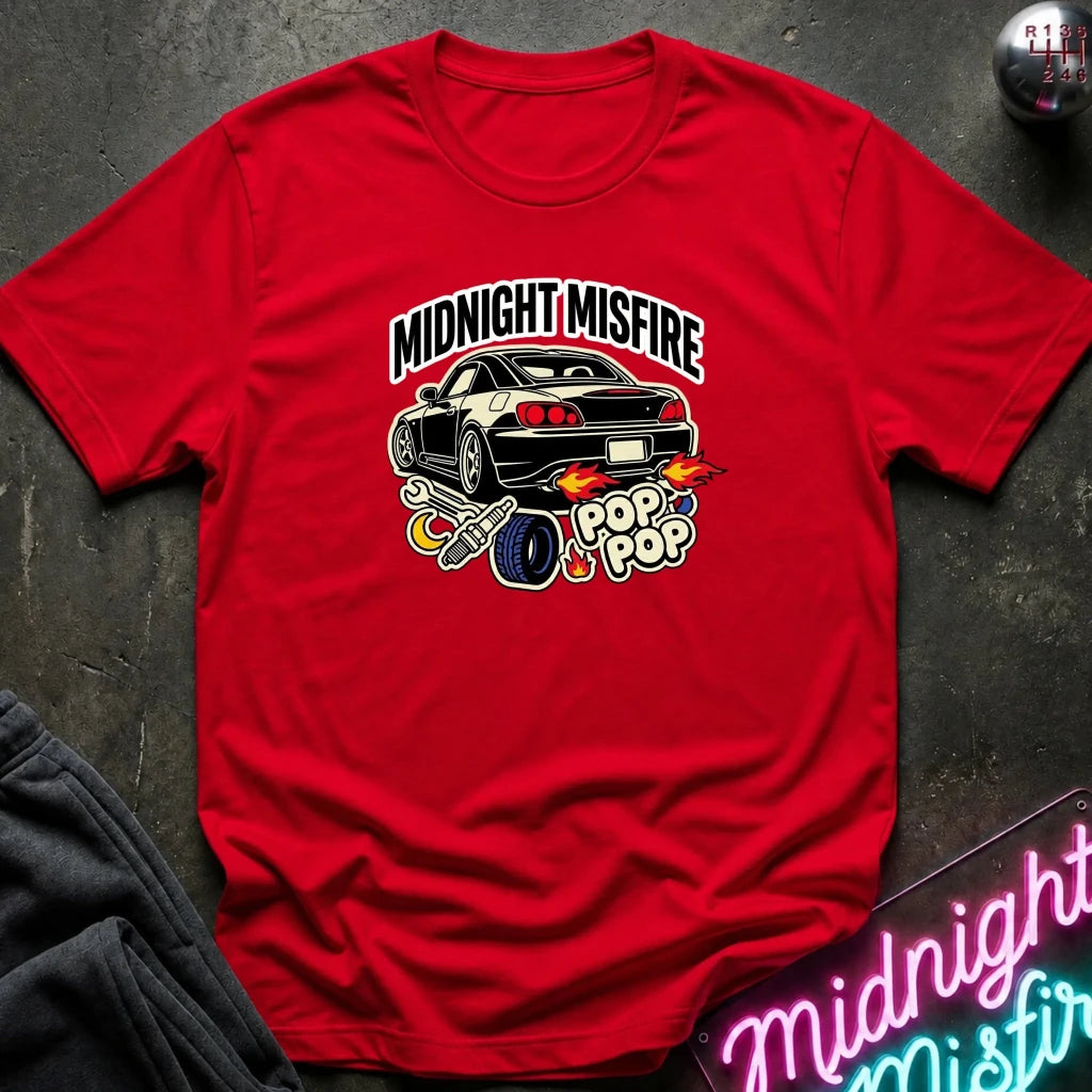 A red short-sleeve graphic t-shirt laid flat on a dark textured surface. The graphic features a black sports car with flames, a blue tire, and the text 'MIDNIGHT MISFIRE' and 'POP POP'.