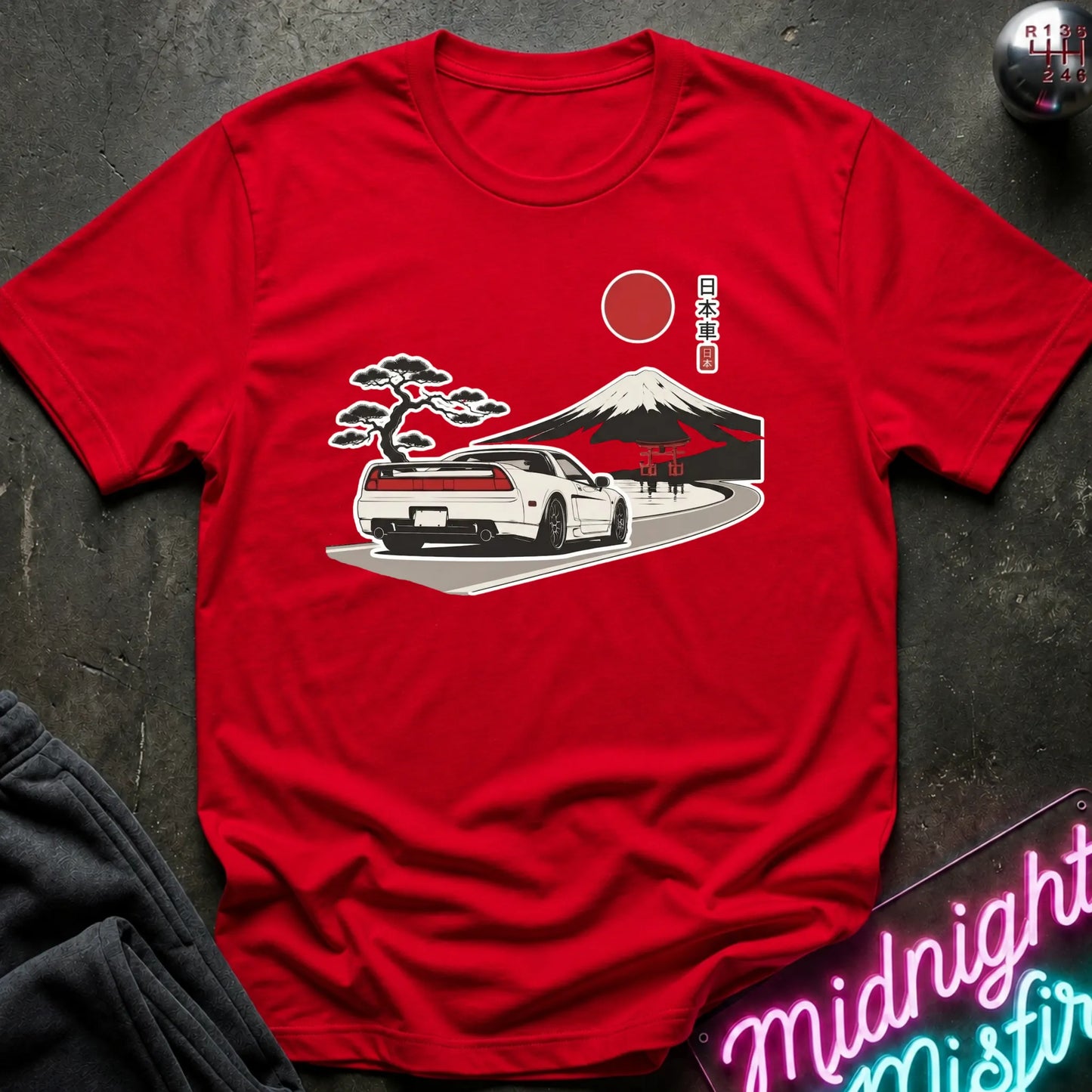 A red short-sleeve crewneck t-shirt laid flat on a dark textured surface, featuring a graphic print of a white sports car, a Japanese mountain landscape, a pine tree, and a red sun with Japanese text.
