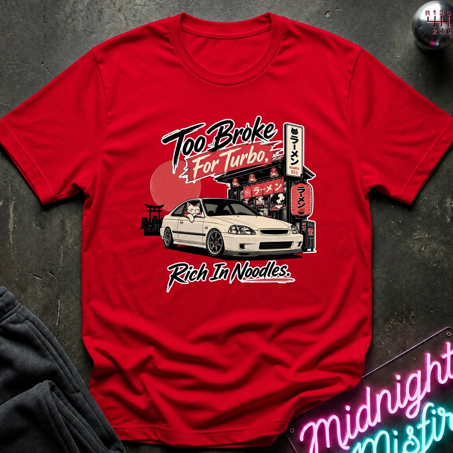 A red short-sleeve graphic T-shirt laid flat on a dark textured surface. The shirt features a printed design of a white sports car in front of a Japanese-style street scene with a red sun and neon signs, plus the text 'Too Broke For Turbo' and 'Rich In Noodles.