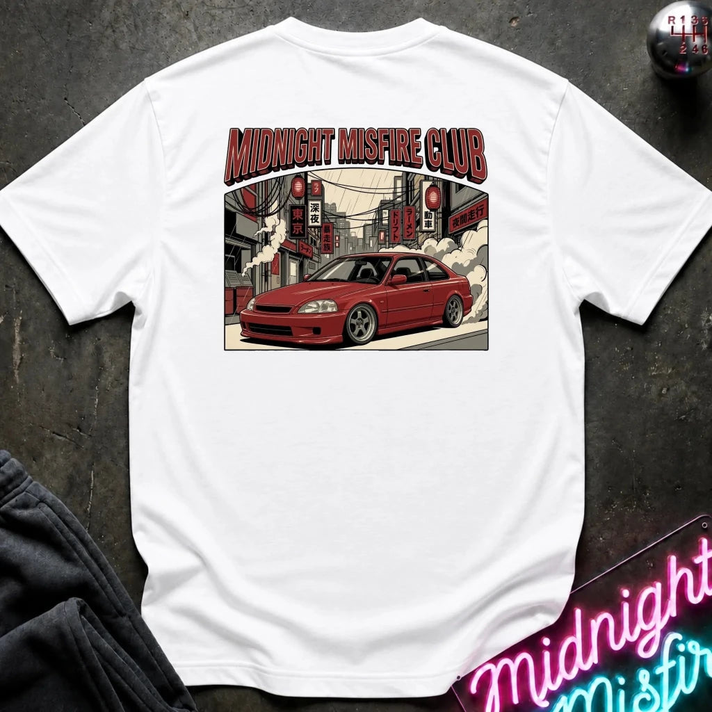 A white short-sleeve t-shirt laid flat on a dark textured surface, featuring a graphic print of a red sports car in a city street with the text 'MIDNIGHT MISFIRE CLUB' above it. A neon sign and dark clothing are partially visible around the edges as styling props.
