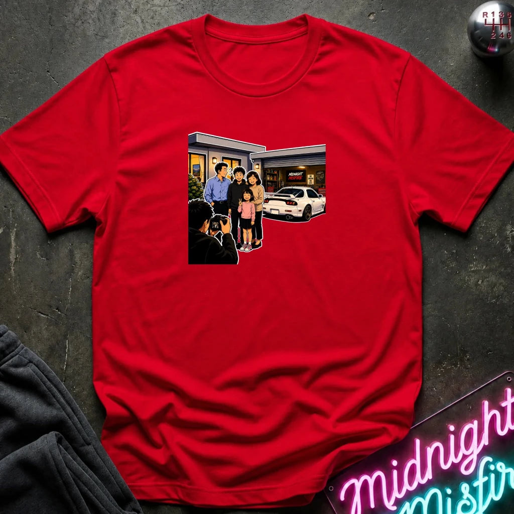 A red short-sleeve crewneck t-shirt laid flat on a dark textured surface, featuring a rectangular graphic print of a family standing outside a building taking a photo.