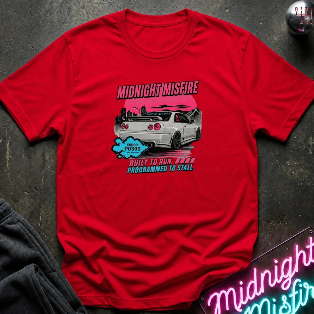 A red short-sleeve graphic t-shirt laid flat on a dark textured surface. The graphic features a silver car against a pink background with the text 'MIDNIGHTMISFIRE' 