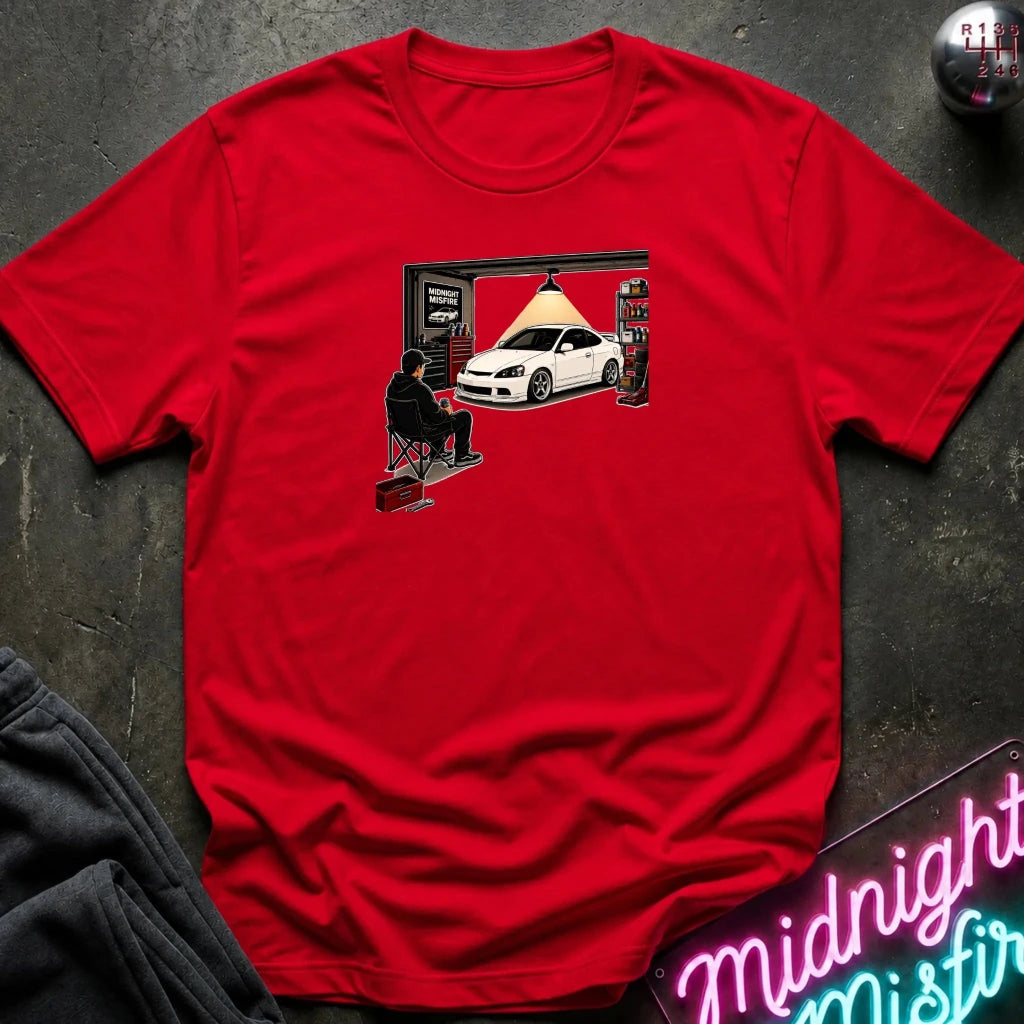 A red short-sleeve crewneck t-shirt featuring a graphic print of a white car inside a garage under a hanging lamp, laid flat on a dark textured surface with a neon sign and dark sweatpants nearby.