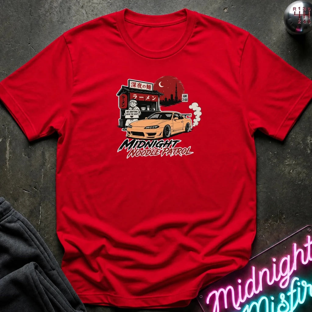 red crewneck t-shirt featuring a graphic print of a yellow Nissan Silvia S15 drift car in front of a red moon and Japanese storefront, with the text 'MIDNIGHT Noodle PATROL'. The shirt is laid flat on a dark textured surface with a neon sign and dark pants partially visible.