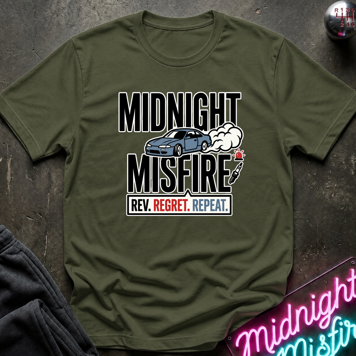 An olive green graphic t-shirt laid flat on a dark textured surface. The shirt features a graphic print with the words 'MIDNIGHT MISFIRE' in bold black letters, a blue car and white smoke