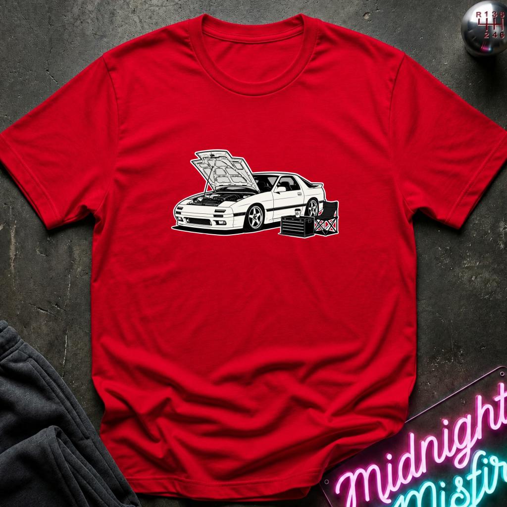 A red short-sleeve crewneck t-shirt featuring a graphic of a white sports car with its hood open, displayed on a dark textured background with a neon sign and sweatpants partially visible.