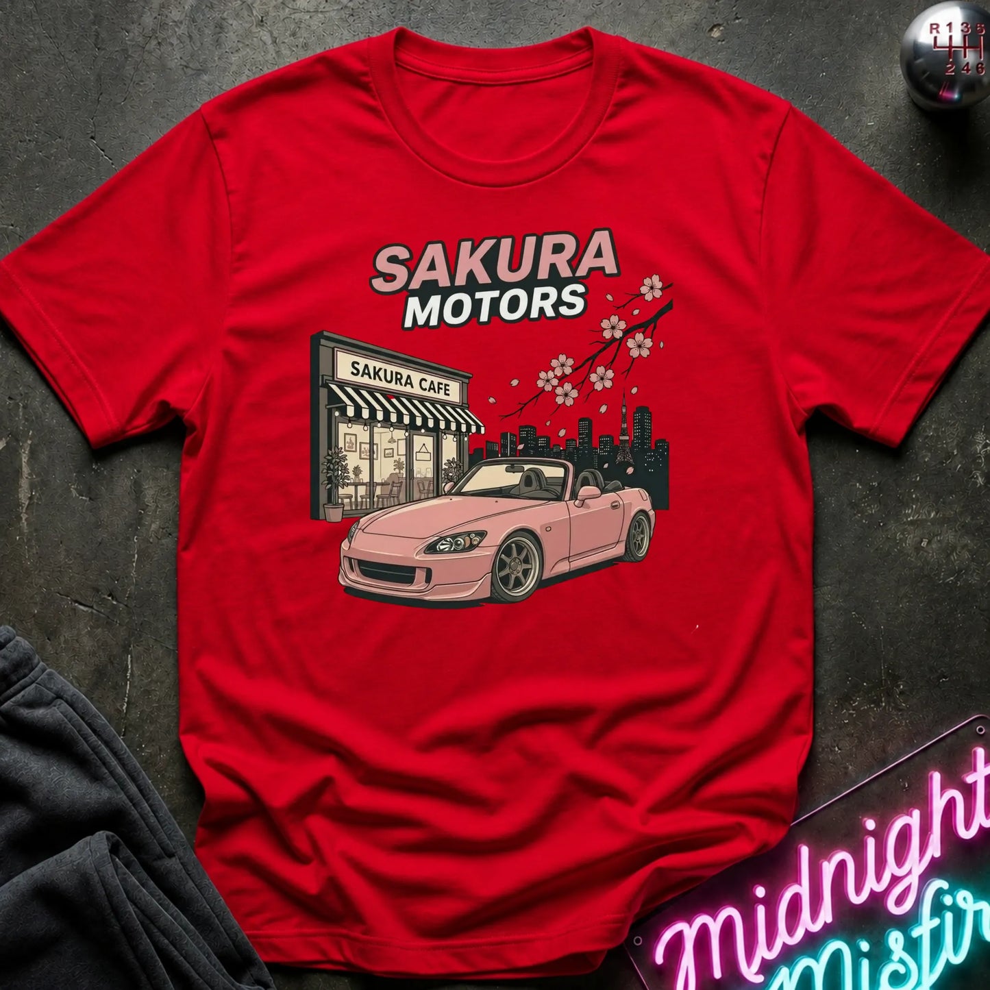 red short-sleeve graphic T-shirt laid flat on a dark textured surface. The shirt features a printed design of a pink convertible sports car in front of a building labeled 'Sakura Cafe', with 'SAKURA MOTORS' text above and cherry blossoms across the top.