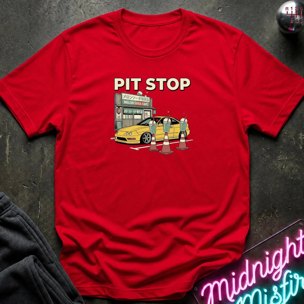 A red short-sleeve crewneck t-shirt laid flat on a dark textured surface. The shirt features a graphic print of a yellow sports car at a pit stop with the text 'PIT STOP' in white.
