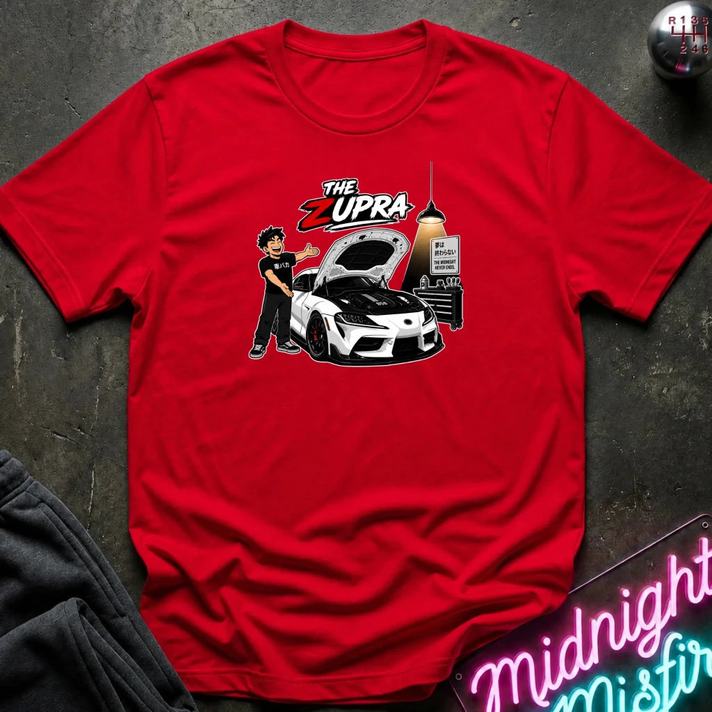 A red short-sleeve graphic t-shirt laid flat on a dark textured surface. The graphic features a cartoon illustration of a man standing next to a white sports car with its hood open, under the text 'THE ZUPRA'.