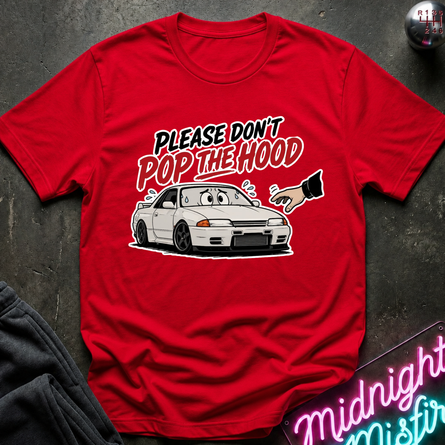 A red short-sleeve graphic T-shirt laid flat on a dark textured surface. The shirt features a cartoon white sports car with a surprised face and the text 'PLEASE DON'T POP THE HOOD' in black and red lettering.