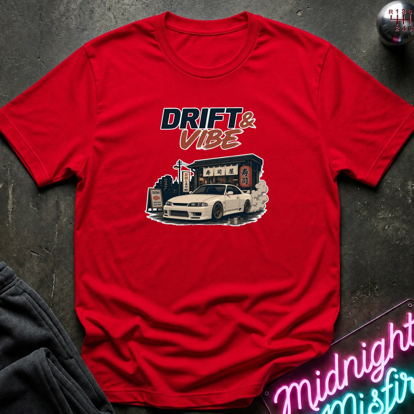 A red short-sleeve graphic t-shirt laid flat on a dark textured surface. The graphic features the text 'DRIFT & VIBE' above an illustration of a white sports car drifting in front of a building.