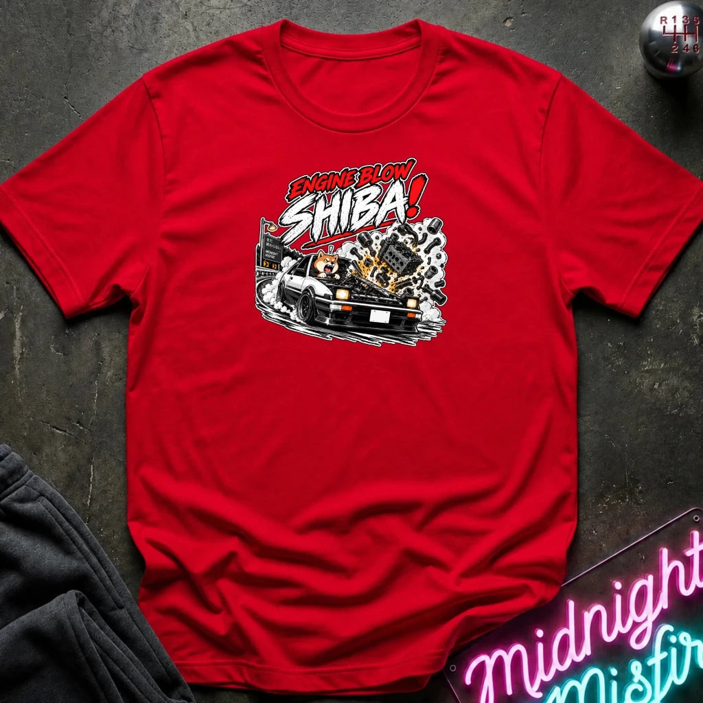 A red short-sleeve graphic t-shirt laid flat on a dark textured surface. The shirt features a stylized graphic of a white sports car with a glowing engine and the text 'ENGINE BLOW SHIBA!' in red, white, and black.