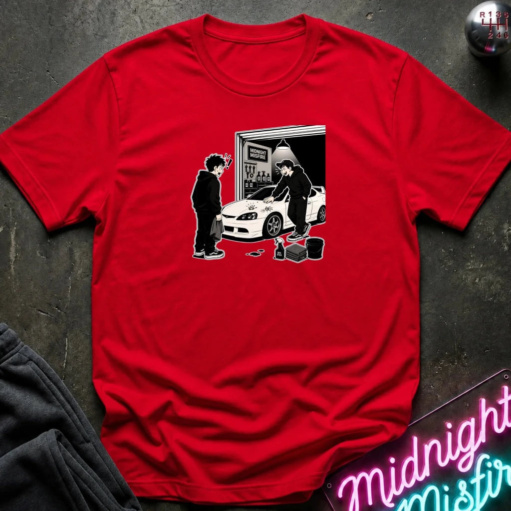 A red short-sleeve crewneck t-shirt with a black and white graphic print of two people standing by a white sports car in front of an open garage door.