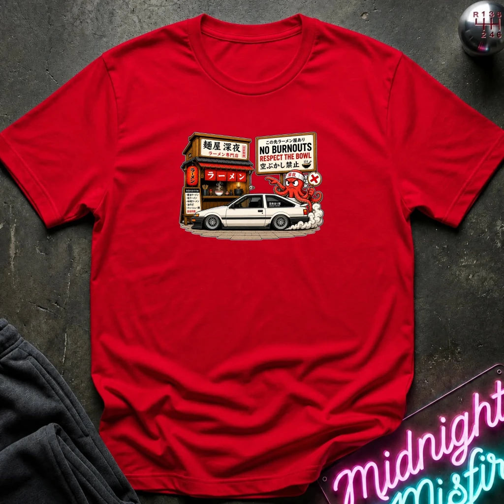 A red short-sleeve crewneck t-shirt laid flat on a dark textured surface, featuring a vintage-style graphic print of a white coupe car parked in front of a Japanese restaurant with a 'No Burnouts' sign.