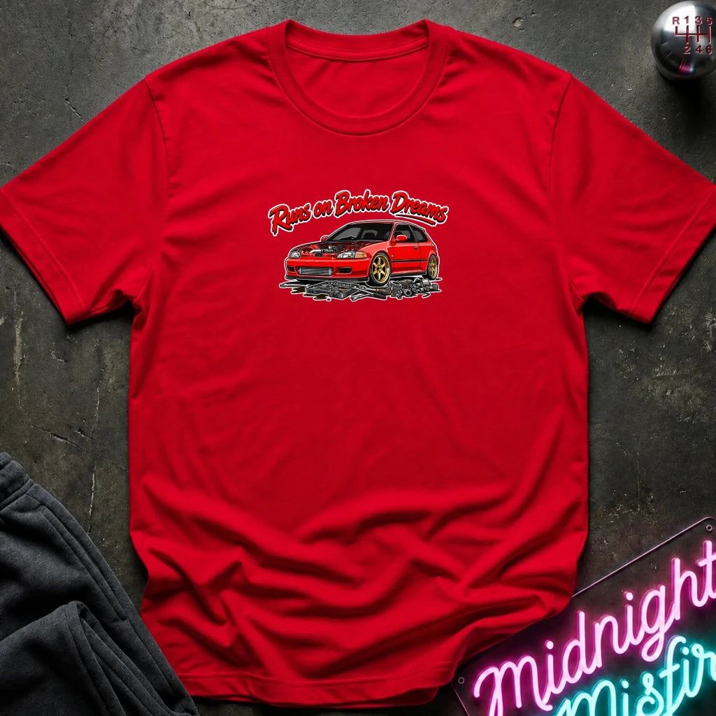A red short-sleeve t-shirt laid flat on a dark textured surface, featuring a graphic of a red sports car with the text 'Ran's Broken Dreams' above it. A gray sweatshirt corner and a neon sign are partially visible as styling props.
