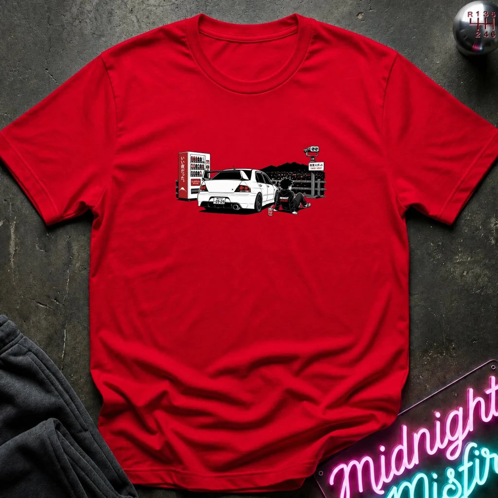 A red short-sleeve t-shirt laid flat on a dark textured surface, featuring a graphic print of a white sports car with a red vending machine.