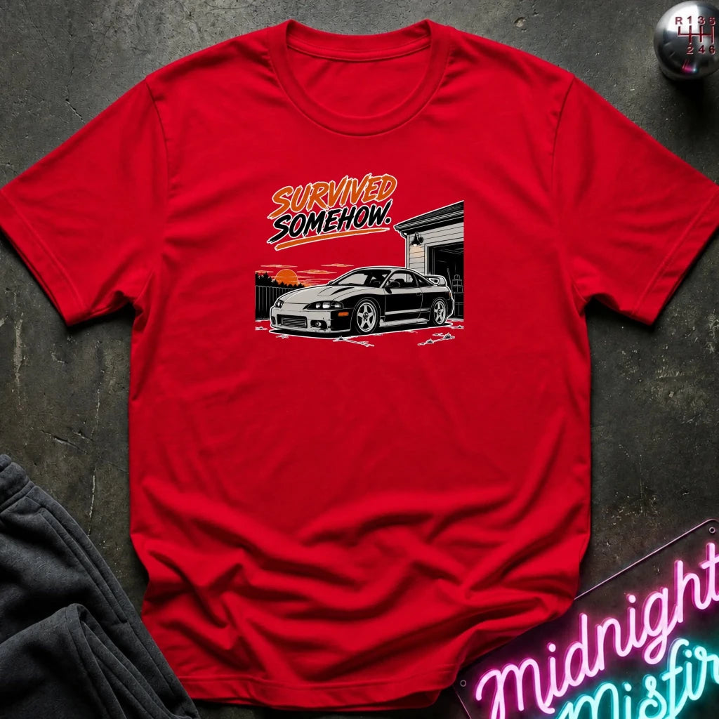 A red short-sleeve graphic t-shirt laid flat on a dark textured surface. The graphic features the text 'SURVIVED SOMEHOW' above a black and white sports car in front of a garage.