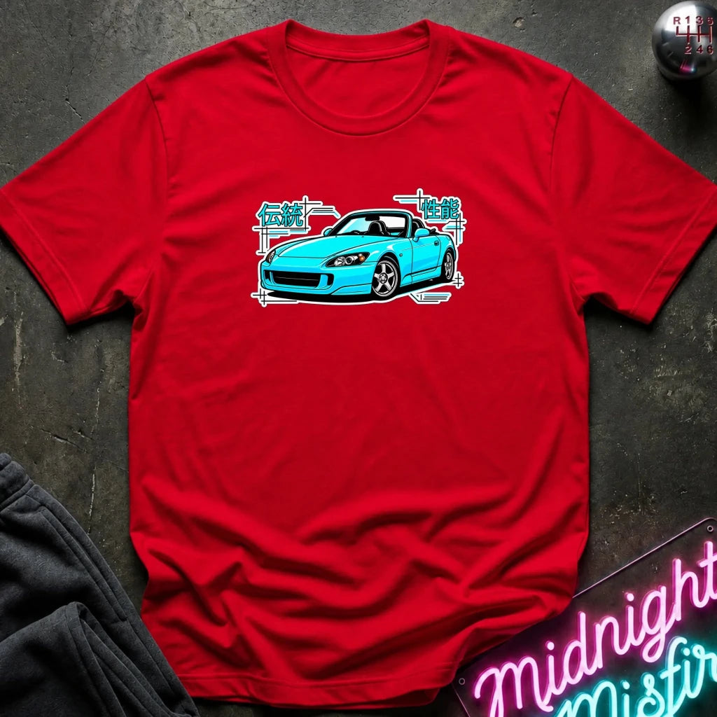 red t-shirt laid flat on a dark textured surface, featuring a graphic print of a bright blue sports car with white and teal text elements. A neon sign and dark clothing are partially visible in the styled flat-lay composition.