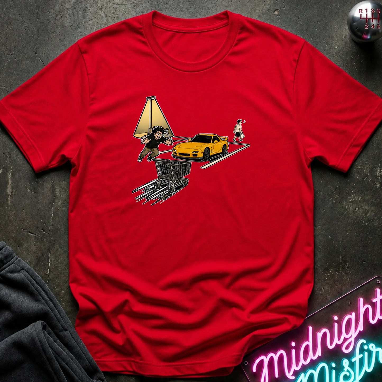 A red short-sleeve crewneck t-shirt laid flat on a dark textured surface, featuring a graphic print of a yellow sports car, a shopping cart, and cartoon characters. 