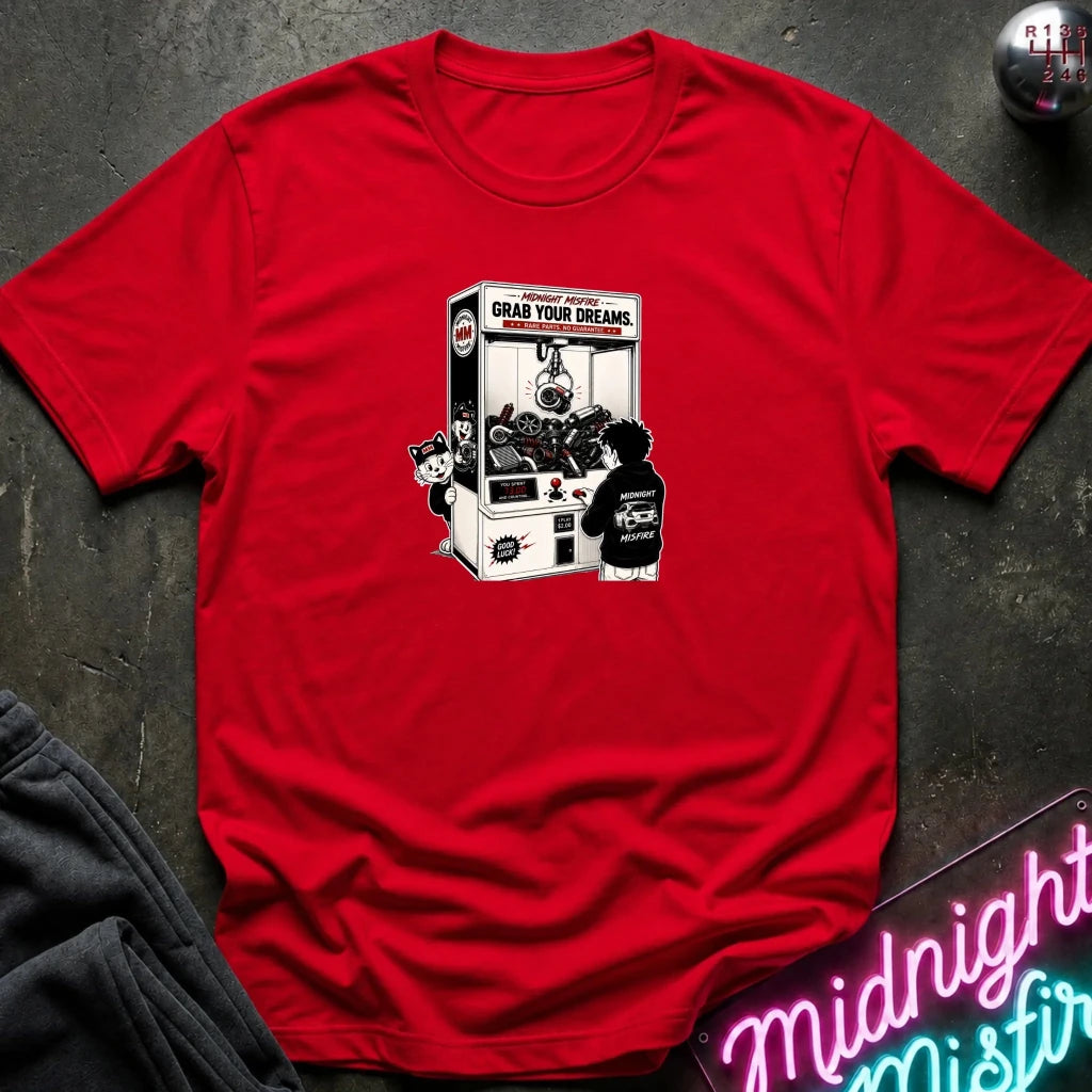 A red short-sleeve crewneck t-shirt laid flat on a dark textured surface, featuring a black and white graphic of a person clawing in a vending machine with the text 'GRAB YOUR DREAMS'.
